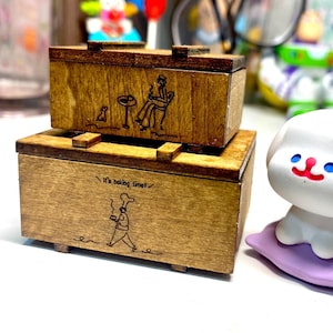 May include: Two wooden boxes stacked on top of each other. The top box has an etched design of a person sitting at a table with a dog. The bottom box has the text "It's baking time!" and an etched chef. The boxes are a light brown color.