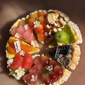Puede incluir: Una tarta de frutas en miniatura y colorida con varios ingredientes, incluyendo rodajas de naranja, uvas y bayas. Cada porción está decorada con pequeñas flores blancas y una etiqueta "Cake". Un postre artesanal encantador.