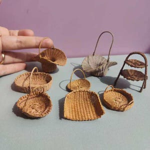 Könnte beinhalten: Eine Sammlung von Miniatur-Weidenkörben in verschiedenen Formen und Größen. Die Körbe sind aus hellbraunem Korbgeflecht gefertigt und umfassen runde, ovale und schaufelförmige Designs. Einige haben Griffe, und einer ist ein zweistöckiges Regal. Die Sammlung ist auf einer hellblauen Oberfläche angeordnet.