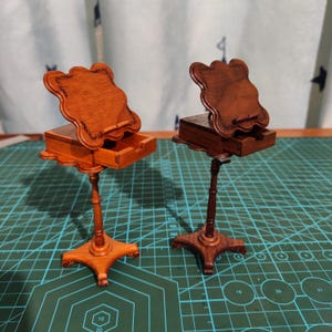 May include: Two wooden music stands with a drawer and a decorative top. The stands are brown and have a carved design. The stands are on a green mat.