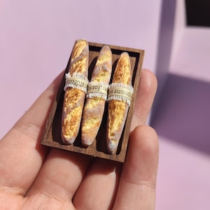 May include: A miniature wooden tray holds three small artisan bread loaves. Each loaf is wrapped with a paper band. The bread has a golden-brown crust and a light interior, suggesting a fresh bake. The tray is held in a hand.