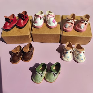 Può includere: Una collezione di scarpe in miniatura in pelle di vari colori, tra cui rosso, bianco, rosa, marrone e verde. Le scarpe, con lacci e dettagli decorativi, sono esposte su piccole scatole di cartone.