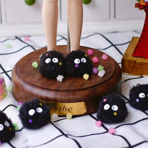 May include: Several small, round, black fuzzy soot sprite figures with large white eyes and tiny colorful star-shaped accents. These plush characters are scattered around a circular wooden display base and a small glass jar filled with colorful star-shaped candies.