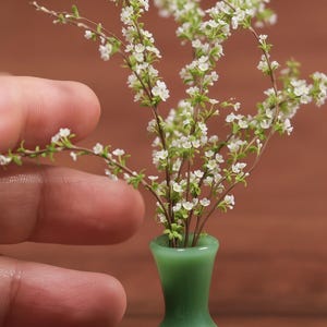 May include: A miniature green vase holds delicate white flowers with green stems and leaves. The vase has a rounded shape and a narrow neck. The flowers are in full bloom, creating a charming, small floral arrangement.