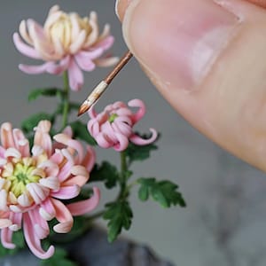 May include: A close-up of a delicate pink and white chrysanthemum flower arrangement. The flowers are made of polymer clay and are being carefully arranged with a small tool.