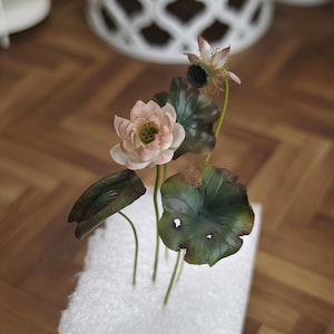 以下が含まれることがあります： 緑の葉が付いた人工のピンクの蓮の花のブーケ。花は白い発泡スチロールの土台に配置されています。