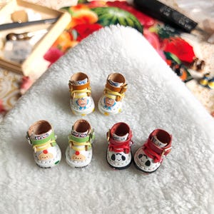 May include: Six pairs of miniature shoes with various designs. Two pairs have yellow and brown accents with cartoon faces. Two pairs have green accents with a dog design. The final two pairs have red accents with a panda design.