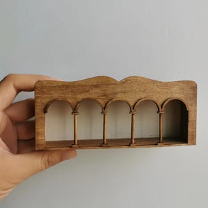 Puede incluir: Una maqueta arquitectónica de madera en miniatura con cinco aberturas arqueadas sostenidas por esbeltas columnas. El modelo tiene un tono de madera cálido y natural y un borde superior curvado. El diseño evoca un estilo clásico o mediterráneo.