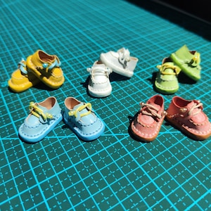 Könnte beinhalten: Eine Sammlung von Miniatur-Lederschuhen in verschiedenen Farben, darunter Gelb, Weiß, Grün, Blau und Rosa. Jedes Paar hat dekorative Schnürsenkel und kleine Perlenakzente. Die Schuhe sind auf einer grünen Rasteroberfläche angeordnet.