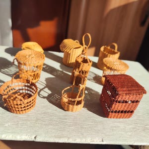 May include: A collection of miniature woven baskets in various shapes and sizes. The baskets are made of natural-colored wicker and range from small, open-topped containers to lidded boxes and handled baskets. The warm tones of the wicker create a rustic, handcrafted aesthetic.