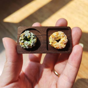 May include: A miniature wooden tray with two small, detailed pastries. One is a ring-shaped cookie covered in almond slices, the other is a similar shape with green and white toppings. The tray is a dark brown color.