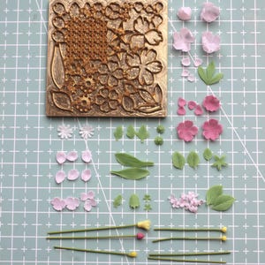 May include: A gold metal floral mould featuring detailed flower and leaf designs. Various pink and white flower petals, green leaves, and stems with yellow and pink buds surround the mould. The items are arranged on a light green grid.