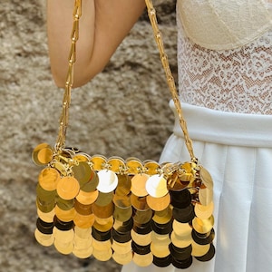 May include: A gold-colored rectangular handbag with a chain strap. The bag is covered in overlapping, circular gold sequins. The chain strap is also gold and appears to be made of interlocking links. The bag is held by a person.