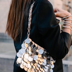 May include: A silver sequined handbag with a silver chain strap. The bag is made of small, round sequins that are arranged in a cascading pattern.