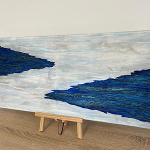 Könnte beinhalten: Abstraktes Kunstwerk auf einer rechteckigen Leinwand, das eine Landschaft in Blau- und Weißtönen darstellt. Die strukturierte Malerei ähnelt Wasser und Himmel. Das Kunstwerk wird auf einer kleinen Holzstaffelei präsentiert.