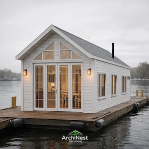 Puede incluir: Una pequeña casa blanca con techo a dos aguas flota sobre el agua. La casa tiene grandes puertas y ventanas de vidrio y está montada sobre un muelle de madera. La casa tiene una chimenea negra y dos luces de pared.