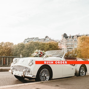 May include: A white vintage convertible car with a floral arrangement on the hood. The car is parked on a street in a city with buildings in the background. The text "RUSH ORDER" is visible on the car.