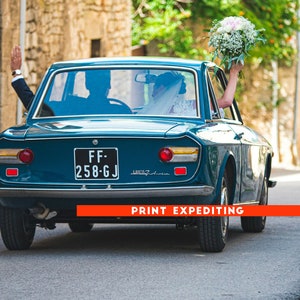May include: A teal blue vintage car with a white license plate that reads "FF-258-GJ" drives down a street. A bride and groom are inside the car, the bride is holding a bouquet of white flowers.