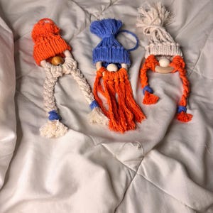 May include: Three handmade gnome ornaments with orange and white yarn beards, white yarn hair, and different colored knit hats. The hats are orange, blue, and white.