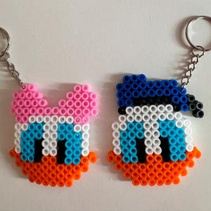May include: Two colourful cartoon character keychains made of perler beads. The keychain on the left is pink, white, blue, and orange and the keychain on the right is blue, white, orange, and black.