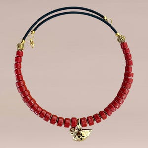 Op de afbeelding: Een rode kralenketting met een goudkleurige hanger. De ketting heeft rechthoekige kralen en een zwart koord. De hanger heeft een bloemenontwerp. De ketting is ongeveer 40 cm lang.