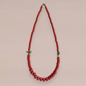 Op de afbeelding: Een rode kralenketting met een kleine goudkleurige vogelbedel. De ketting heeft rechthoekige kralen en een sluiting aan de bovenkant. Er zijn ook een paar blauwe en gele kralen aanwezig. De ketting wordt gepresenteerd tegen een lichtroze achtergrond.