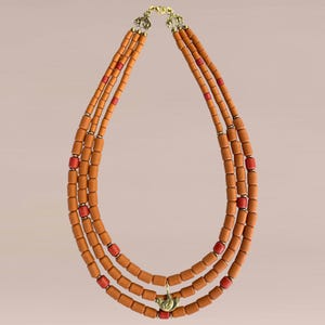Puede incluir: Collar de tres hilos con cuentas cilíndricas de color naranja-marrón y detalles rojos. El collar tiene un cierre dorado y un pequeño colgante dorado. Las cuentas están dispuestas en un diseño en capas.
