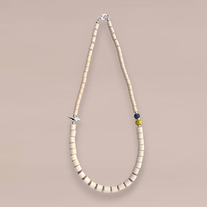 Op de afbeelding: Een crèmekleurige kralenketting met een zilveren vogelbedel. De ketting heeft cilindrische kralen en een sluiting. Een paar blauwe en gele kralen bevinden zich bij de sluiting. De ketting wordt getoond tegen een lichtroze achtergrond.