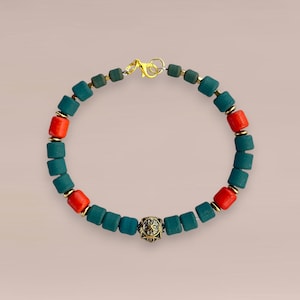 Op de afbeelding: Een armband met vierkante teal en rode kralen, afgewisseld met goudkleurige accenten. De armband heeft een goudkleurige sluiting en een decoratieve goudkleurige kraal in het midden. Het is een sieraad.