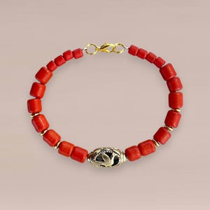 Op de afbeelding: Rode koraal kralenarmband met goudkleurige accenten en sluiting. De armband heeft rechthoekige koraalkralen en een decoratief goudkleurig middelpunt met een vogelontwerp. Een stijlvol sieraad.