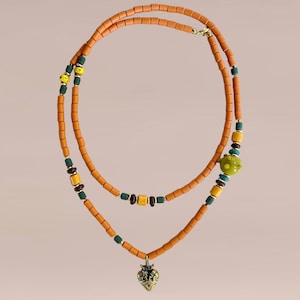 Op de afbeelding: Een dubbele ketting met oranje cilindrische kralen. Het heeft groene, gele en bruine accentkralen en een goudkleurige hanger. De ketting wordt gesloten met een gouden sluiting.