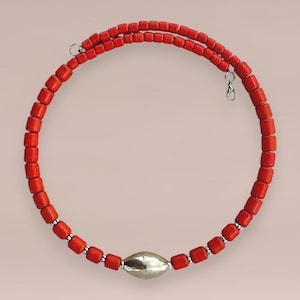 Op de afbeelding: Een rode kralenketting met een zilveren sluiting en een grote zilveren ovaalvormige kraal in het midden. De ketting is gemaakt van kleine, vierkante rode kralen. De ketting is ongeveer 40 cm lang.