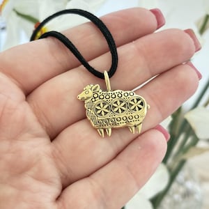 May include: A gold-coloured sheep pendant on a black cord necklace. The sheep pendant has detailed floral patterns on its body and is suspended from a small loop. The necklace is shown against a soft background.