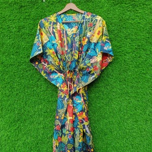 May include: A long, flowing kaftan with a vibrant, colorful print. The fabric features a dark blue background with a tropical design of flowers, trees, and figures in shades of yellow, red, and blue. The kaftan has a V-neck and a drawstring waist.