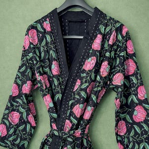 Könnte beinhalten: Ein schwarzer Bademantel mit einem Blumenmuster aus rosa Blüten und grünen Blättern. Der Bademantel hat ein Wickeldesign mit passendem Gürtel. Die Kanten sind mit einem kontrastierenden schwarzen Stoff und weißen Nähten versehen.