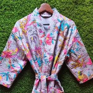 Könnte beinhalten: Grauer, gesteppter Bademantel mit einem lebendigen Blumen- und Vogelmuster in Pink, Gelb, Blau und Lila. Der Bademantel hat einen passenden Gürtel und zwei Vordertaschen. Der Bademantel hängt an einem Holzbügel vor grünem Hintergrund.