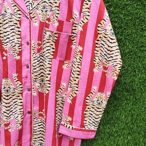 May include: Pink and red striped pajama top with a tiger print. The long-sleeved top has a button-down front, a chest pocket, and white trim. The tiger print features tan and black stripes.