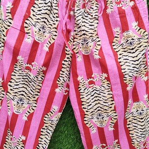 May include: Pink and red striped pajama pants with a repeating tiger print. The pants have a drawstring waist and a relaxed fit. The tigers are tan with black stripes.
