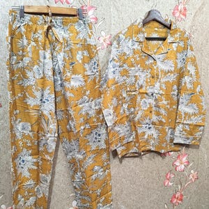 May include: Mustard yellow pajama set with a floral pattern in white and blue. The set includes a button-down shirt and drawstring pants, both hanging on separate hangers. The pajamas have white trim.
