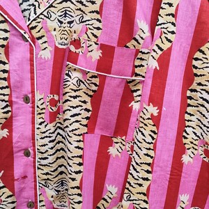 May include: Pink and red striped pajama shirt with a tiger print. The shirt has a button-down front, a chest pocket, and white trim. The tiger print features tan and black stripes.