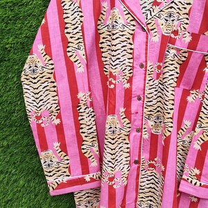 May include: Pink and red striped pajama set with a tiger print. The long-sleeved top and pants feature a repeating pattern of tigers with black and tan stripes. White trim accents the collar, cuffs, and pocket.