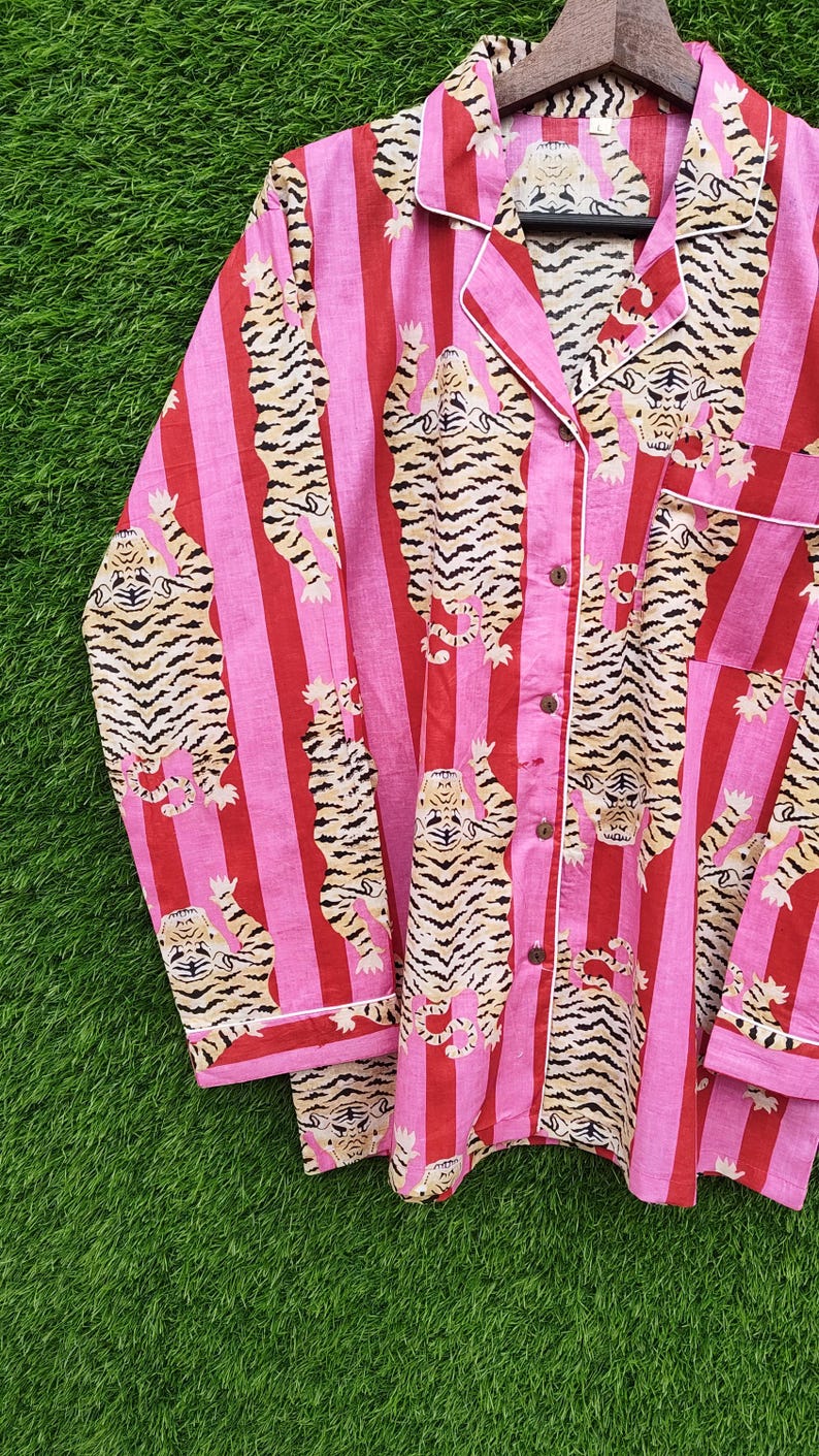 May include: Pink and red striped pajama set with a tiger print. The long-sleeved top and pants feature a repeating pattern of tigers with black and tan stripes. White trim accents the collar, cuffs, and pocket.