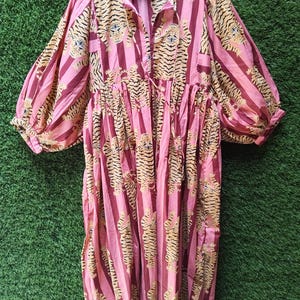 Könnte beinhalten: Ein knielanges Kleid mit einem sich wiederholenden Tigermuster auf vertikalen Streifen in Rosa- und Dunkelrottönen. Das Kleidungsstück hat lange, voluminöse Puffärmel mit gerafften Bündchen, einen geschlitzten Ausschnitt mit Bindebändern und eine geraffte Taille.