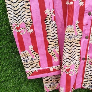 May include: Pink and red striped pajama set with a repeating tiger print. The tigers are tan and black, and the pajamas have white trim and wooden buttons. The fabric is soft and lightweight, perfect for lounging.