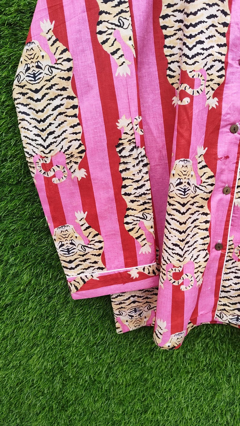May include: Pink and red striped pajama set with a repeating tiger print. The tigers are tan and black, and the pajamas have white trim and wooden buttons. The fabric is soft and lightweight, perfect for lounging.