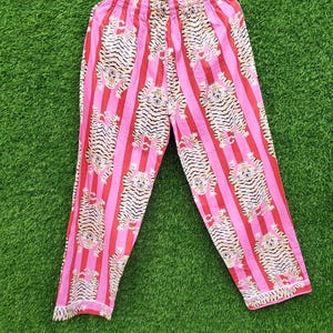 May include: Pink and red striped pajama pants with a repeating tiger print. The pants are made of a lightweight fabric and have a drawstring waist. The pants are hanging on a wooden hanger against a green background.
