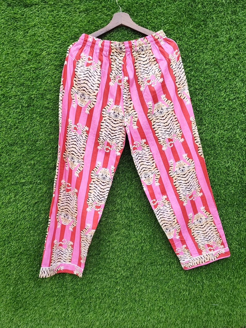 May include: Pink and red striped pajama pants with a repeating tiger print. The pants are made of a lightweight fabric and have a drawstring waist. The pants are hanging on a wooden hanger against a green background.