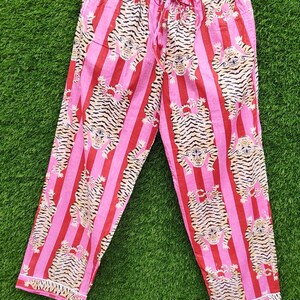 May include: Pink and red striped pajama pants with a tiger print. The pants are hanging on a wooden hanger against a green background. The pajama pants have a drawstring waist and white trim.