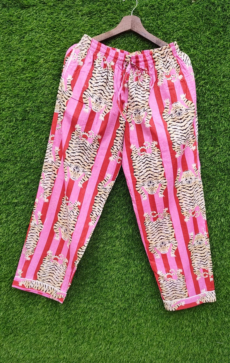 May include: Pink and red striped pajama pants with a tiger print. The pants are hanging on a wooden hanger against a green background. The pajama pants have a drawstring waist and white trim.