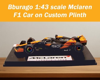 May include: A 1:43 scale model McLaren F1 car, in black and orange, presented on a custom black plinth. The plinth displays the McLaren Formula 1 Team logo. The car has multi-colored wheels and the word "Chrome" is visible.